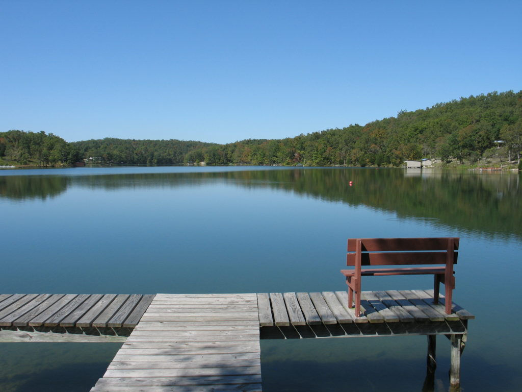 A simple fishing dock with a red wooden bench sits above the clear waters of Lake Vagabond in Ozark Acres